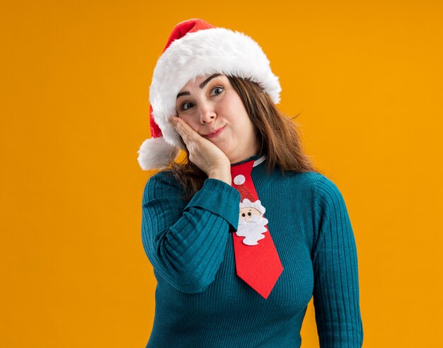 Excited adult caucasian woman with santa hat and santa tie puts hand on face and looks at camera isolated on orange background with copy space