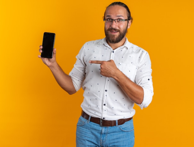 Excited adult handsome man wearing glasses showing mobile phone pointing at it looking at camera isolated on orange wall