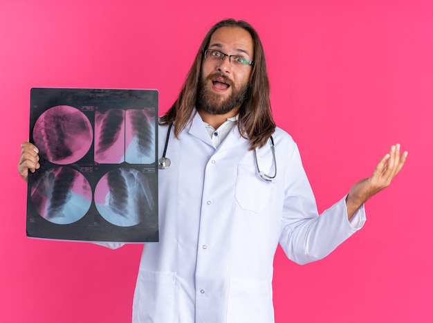Взволнованный взрослый мужчина-врач в медицинском халате и стетоскоп в очках смотрит в камеру, показывающую рентгеновский снимок и пустую руку, изолированную на розовой стене
