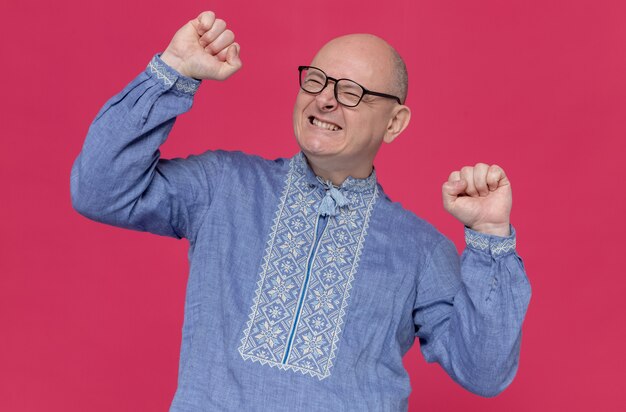 Excited adult man in blue shirt wearing glasses raising fists up 