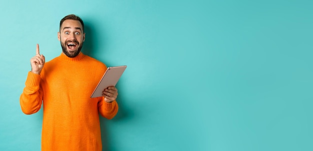 Excited adult man using digital tablet and having an idea raising finger eureka sign standing over l