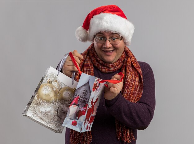 Excited adult man wearing glasses and santa hat with scarf around neck holding christmas gift bags opening one looking inside it isolated on white wall