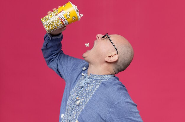 Excited adult slavic man in blue shirt wearing optical glasses holding and eating popcorn bucket 