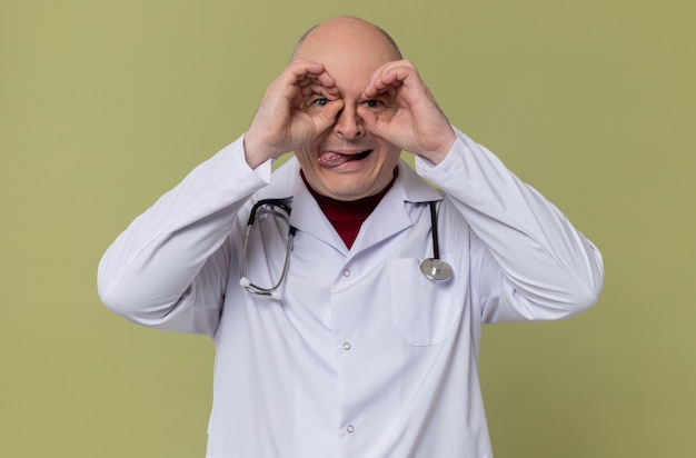 Возбужденный взрослый славянский мужчина в медицинской форме со стетоскопом высунул язык и сквозь пальцы
