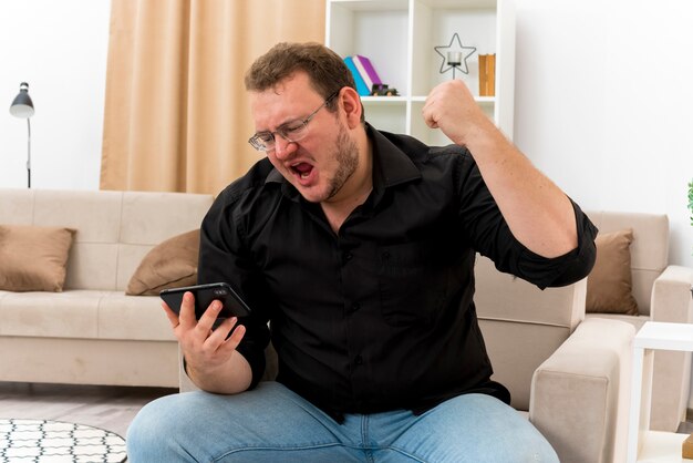 Возбужденный взрослый славянский мужчина в оптических очках сидит на кресле с поднятым кулаком и смотрит на телефон в гостиной