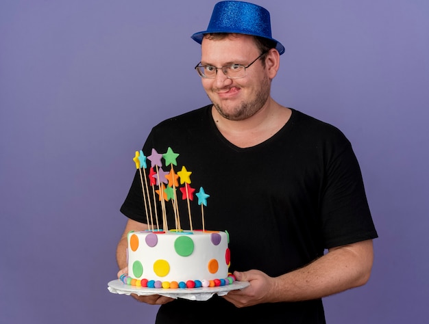 Excited adult slavic man in optical glasses wearing blue party hat stucks out tongue holding birthday cake 