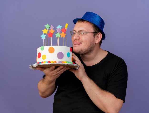 Excited adult slavic man in optical glasses wearing blue party hat stucks out tongue and holds birthday cake 