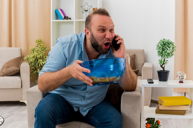 Excited adult slavic man sits on armchair talking on phone and holding bowl of chips inside the living room