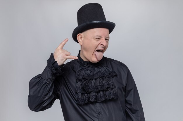 Excited adult slavic man with top hat and in black gothic shirt stucks out tongue and gestures horns sign