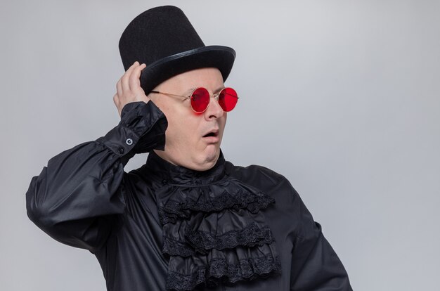 Excited adult slavic man with top hat and with sunglasses in black gothic shirt looking at side 