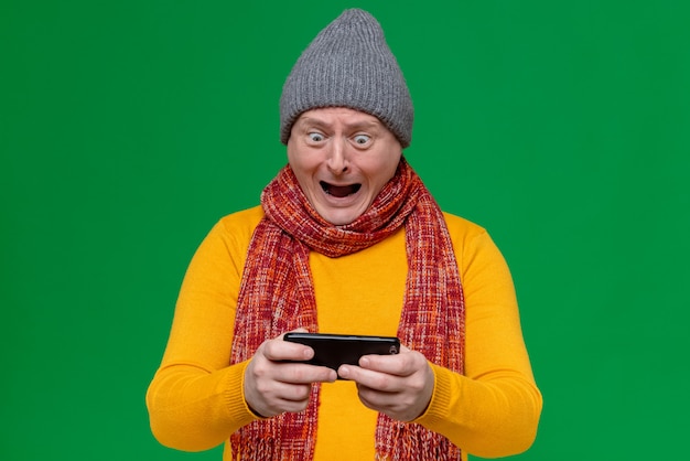 Excited adult slavic man with winter hat and scarf around his neck holding and looking at phone