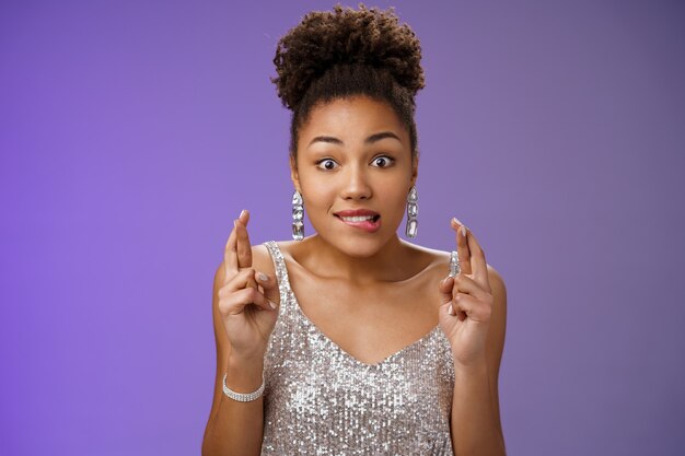 Excited african-american girl excited eager win cross fingers good luck biting lip desire anticipation making wish, eager receive gift, standing optimistic thrilled in silver dress blue background.