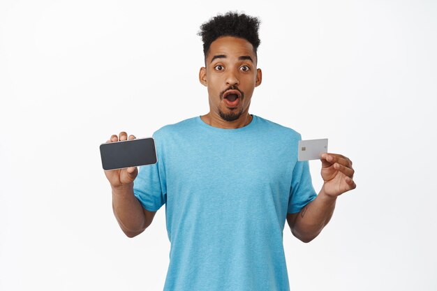 Excited african american man gasp amazed, say wow, showing horizontal smartphone screen and credit card, showing mobile phone application, shopping app, standing against white background