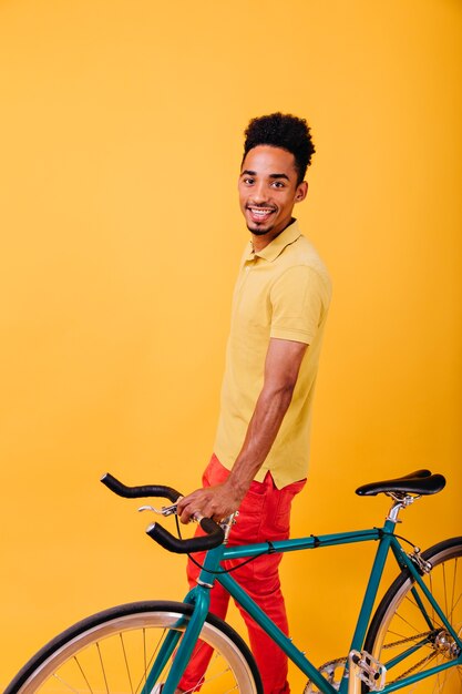 緑の自転車でポーズをとってスタイリッシュな服を着て興奮したアフリカ人。自転車で楽しんでいる笑顔の黒人男性の屋内写真。