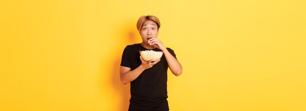 Excited and amazed asian guy watching interesting movie and eating popcorn from bowl standing yellow