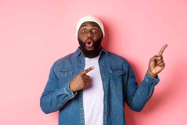 Excited and amazed Black man showing awesome offer, pointing fingers right at copy space, standing in hipster beanie and denim shirt on pink background