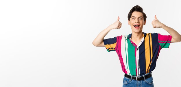 Excited and amazed young man showing thumbs up with impressed expression standing over white background