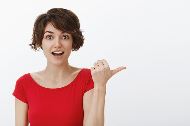 Excited and amused woman pointing thumb right at your logo or advertisement