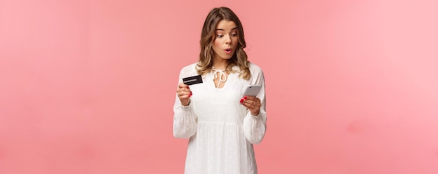 興奮して興味をそそられる白いドレスを着た若いブロンドの女の子が急いでオンラインショッピングサイトで注文し、割引が終了する前に携帯電話のピンクの背景にクレジットカードの数字を入力します
