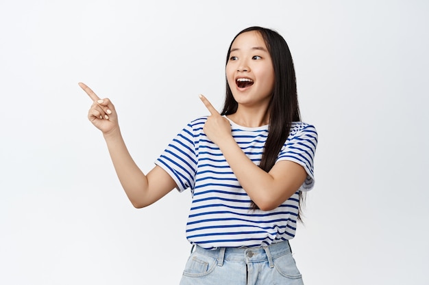 Excited asian girl gasping in awe, pointing fingers and looking left with amazed and surprised face expression, standing on white.