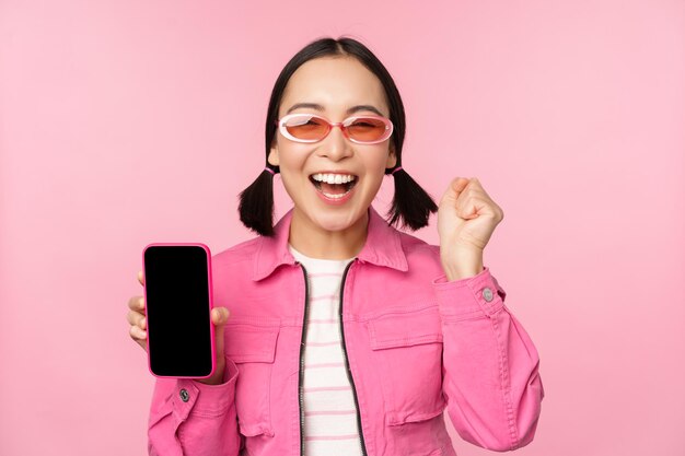 흥분한 아시아 소녀가 웃고 분홍색 배경 위에 서 있는 휴대폰 화면 스마트폰 애플리케이션을 보여줍니다.