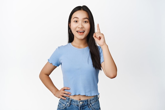Excited asian girl pitching an idea raising finger up and showing big news making announcement standing over white background