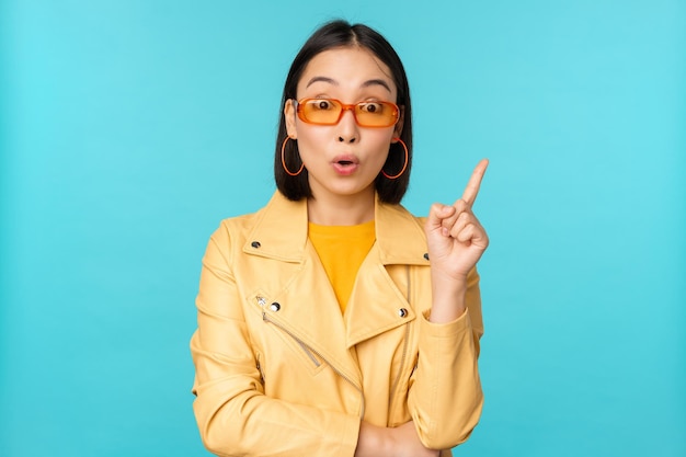 Excited asian girl raising finger suggesting smth eureka sign has plan standing in sunglasses over blue background