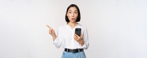 携帯電話を保持し、白い背景にスマートフォンで立っているプロモーションテキストを示す空のコピースペースに人差し指を指してモバイルアプリで買い物をする興奮したアジアの女の子