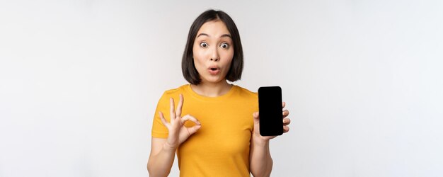 Excited asian girl showing mobile phone screen okay sign recommending smartphone app standing in yel