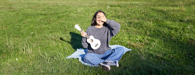 excited asian girl sits in park with ukulele plays instrument and feels happy upbeat positive