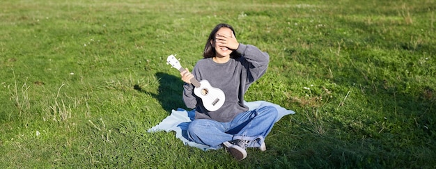 Excited asian girl sits in park with ukulele plays instrument and feels happy upbeat positive