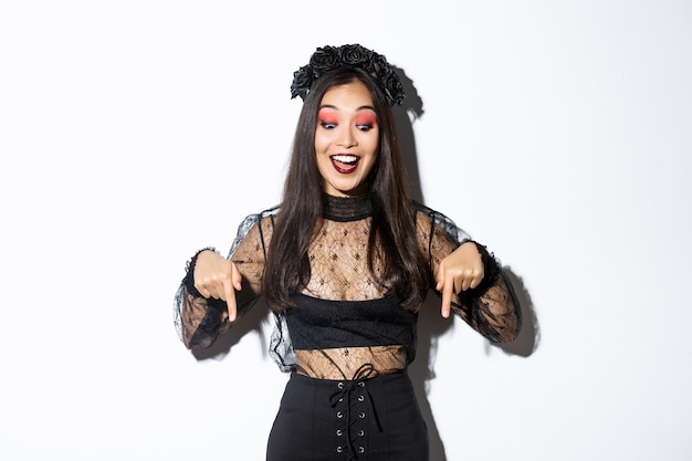 Excited asian woman dressed-up for halloween, smiling amused and pointing fingers down, showing advertisement, standing in party costume over white background.