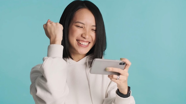 Excited Asian woman emotionally playing on mobile phone celebrating win in online game isolated on blue background