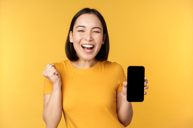 Excited asian woman showing smartphone app and triumphing celebrating on mobile phone standing over yellow background