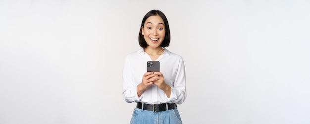 Возбужденная азиатка улыбается, реагируя на информацию по мобильному телефону, держа смартфон и счастливо глядя на