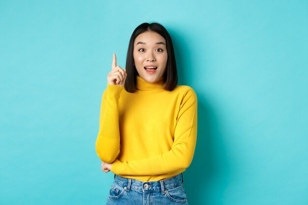 Excited asian woman with short dark hair, pitching an idea, raising finger in eureka gesture and smiling, standing over blue background