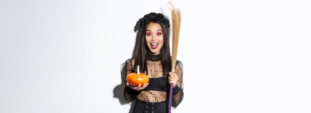 Ragazza asiatica attraente emozionante che celebra halloween che indossa il costume della strega che tiene zucca e scopa