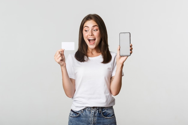 Excited attractive girl showing smartphone screen and credit card, white.