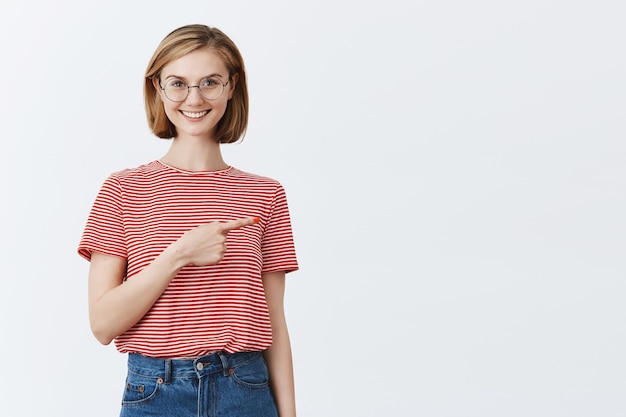 Excited attractive young girl in glasses showing way, pointing finger right at copyspace