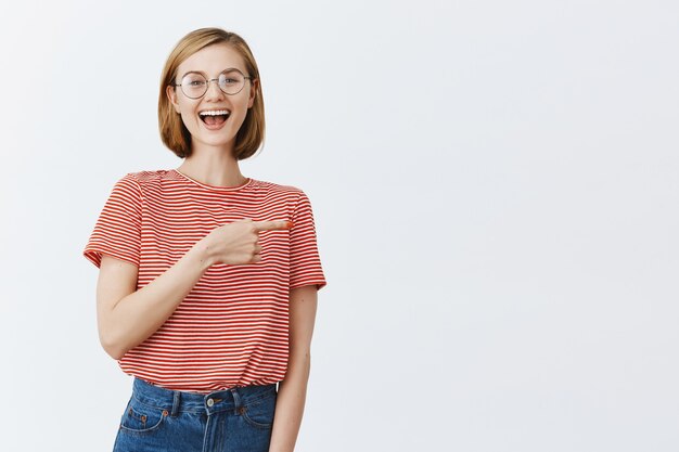 Excited attractive young girl in glasses showing way, pointing finger right at copyspace