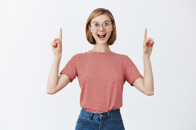 Excited attractive young girl in glasses showing way, pointing fingers up at copyspace