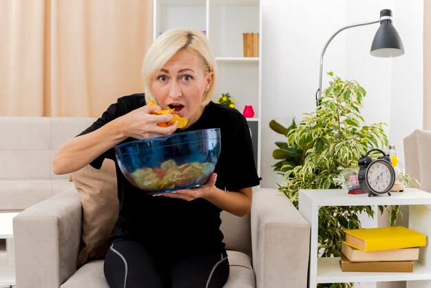 흥분된 아름 다운 금발 러시아 여자는 거실 내부에 칩 그릇을 들고 먹는 안락의 자에 앉아