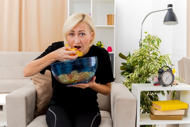 Бесплатное фото Возбужденная красивая русская блондинка сидит на кресле и ест миску чипсов в гостиной