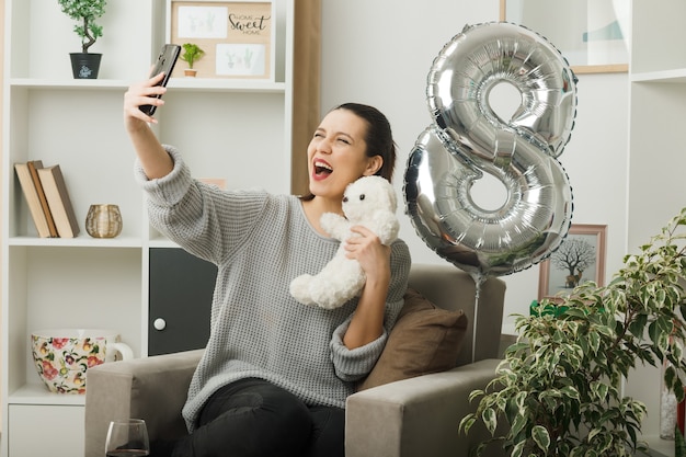 Excited beautiful girl on happy women day holding teddy bear take a selfie sitting on armchair in living room