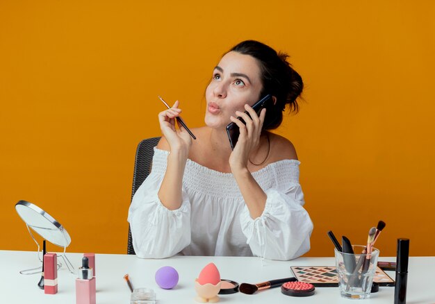 Excited beautiful girl sits at table with makeup tools talks on phone holding makeup brush looking up isolated on orange wall