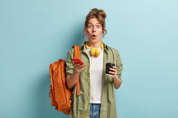 Excited beautiful hipster girl opens mouth from surprisement, reads news in internet, uses modern cell phone and headphones for listening music or audio book, holds takeaway coffee, going on classes