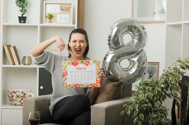 Excited beautiful woman on happy women day holding and points at calendar sitting on armchair in living room