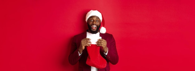 Uomo nero eccitato calza di natale aperta con regali e dolci sorridendo felice in piedi in cappello di babbo natale a