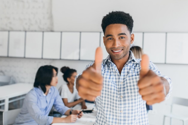 Excited black manager showing thumbs up after successul negotiation with partners. Indoor portrait of asian employees with african worker in blue shirt on foreground.