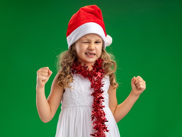 Excited blinked little girl wearing christmas hat with garland on neck showing yes gesture isolated on green wall